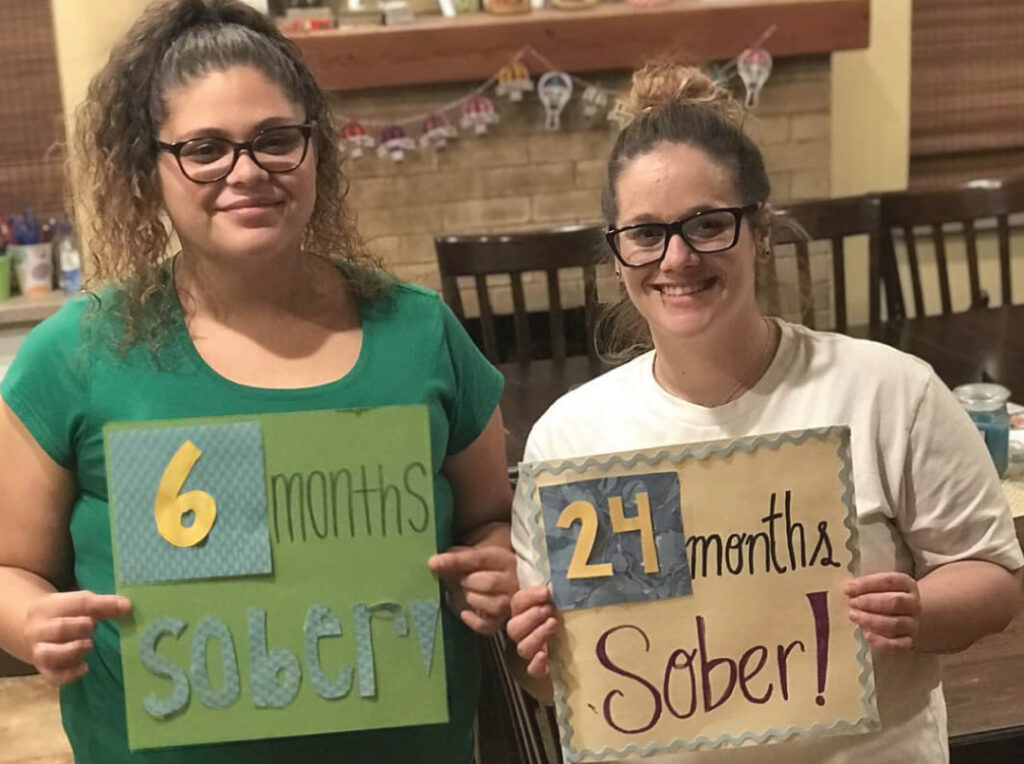 Image of two women smiling, holding up signs indicating months of sobriety